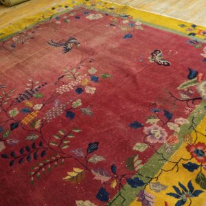 A distressed rug with a red field and yellow border featuring colorful bird and floral patterns on wood floors.