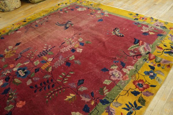 A distressed rug with a red field and yellow border featuring colorful bird and floral patterns on wood floors.
