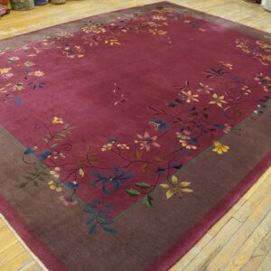 Berry-colored rug with flowing floral pattern and brown border is laid over worn light wood floors.