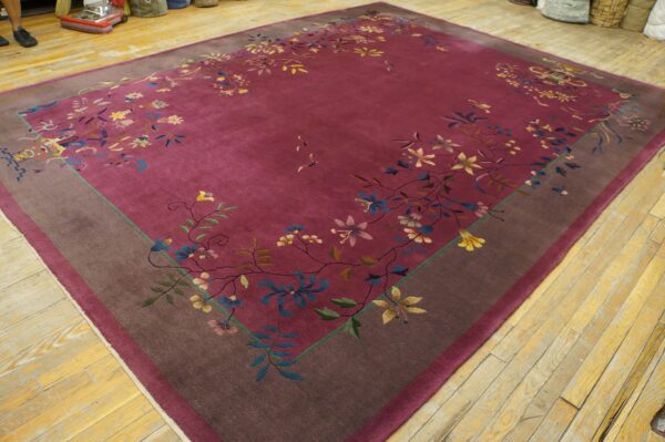 Berry-colored rug with flowing floral pattern and brown border is laid over worn light wood floors.