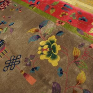 Plush brown rug with multicolored floral and knot motifs, framed by a red border next to wood plank flooring.