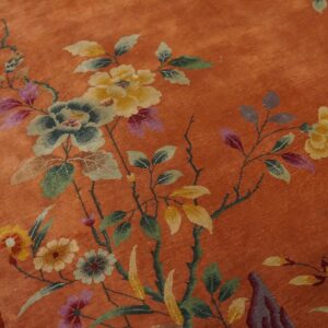 Burnt orange rug displaying a detailed floral pattern featuring yellow, faded green, and purple blooms beside worn wood flooring.