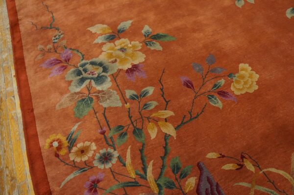 Burnt orange rug displaying a detailed floral pattern featuring yellow, faded green, and purple blooms beside worn wood flooring.