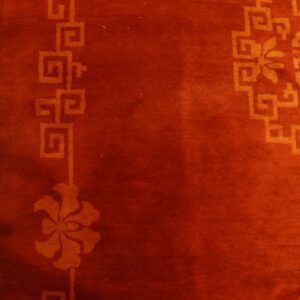 Deep red rug featuring orange fretwork and small blue floral details rests beside worn yellow wood flooring.