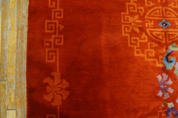 Deep red rug featuring orange fretwork and small blue floral details rests beside worn yellow wood flooring.