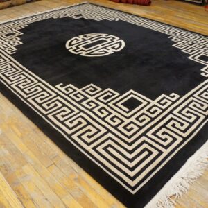 A large black rug with a cream geometric meander border and central medallion lies on distressed hardwood floors.