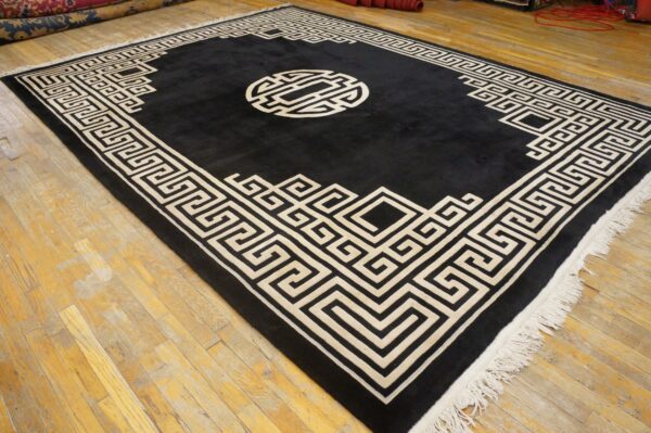 A large black rug with a cream geometric meander border and central medallion lies on distressed hardwood floors.