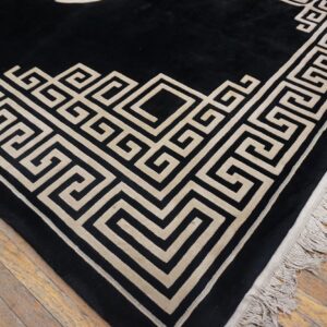Black rug with a light tan geometric Greek key border and fringes on a wooden floor.