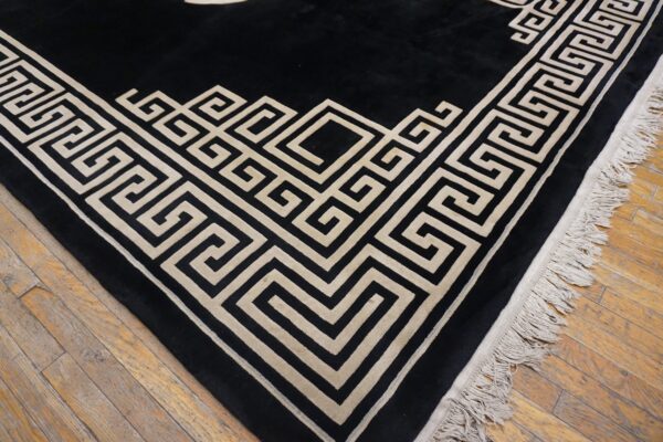Black rug with a light tan geometric greek key border and fringes on a wooden floor.