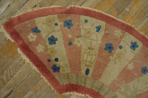 A fan-shaped rug in pink and beige with blue floral motifs rests on distressed wood planks.