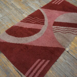 High-pile rectangular rug with red, pink, and taupe geometric shapes on distressed wood flooring.