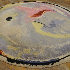 Round, thick pile abstract rug in muted gray, pink, yellow, and black lies on distressed wood planks.