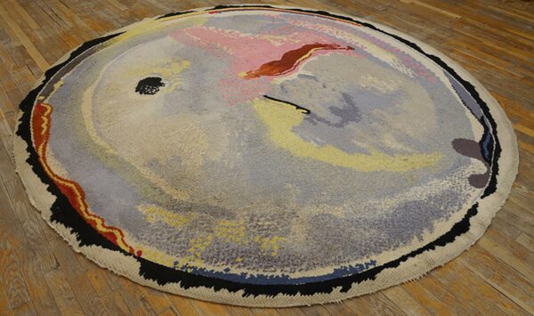 Round, thick pile abstract rug in muted gray, pink, yellow, and black lies on distressed wood planks.