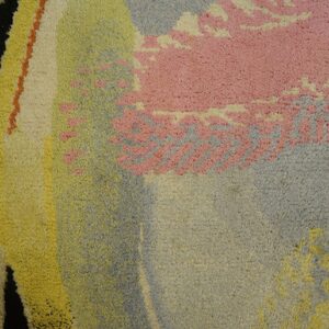 Close-up of a high-pile rug featuring large abstract forms in pink, blue, yellow, and black over wood planks.