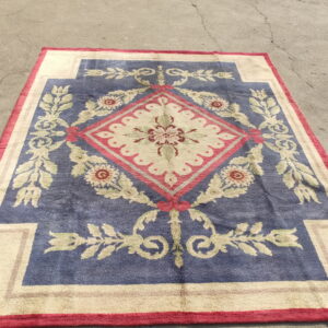 Ornate blue and cream rug with a red-outlined floral medallion rests on gray, cracked pavement.