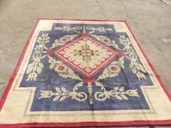 Ornate blue and cream rug with a red-outlined floral medallion rests on gray, cracked pavement.
