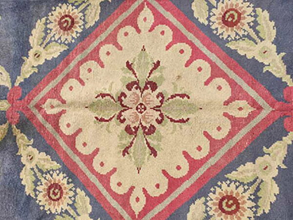 Blue rug detail showing a cream scalloped diamond surrounded by red, sage, and burgundy floral garlands.