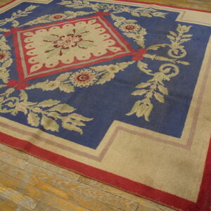 Blue rug with beige floral and scroll patterns and red accents on distressed wooden plank flooring.