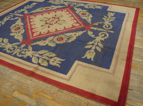 Blue rug with beige floral and scroll patterns and red accents on distressed wooden plank flooring.