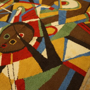 Abstract high-pile rug in brown, yellow, red, blue, and green with a central mask motif on wood floors.