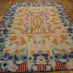 Abstract high-pile rug features pink, orange, and blue geometric patterns with a stylized floral border on wood planks.