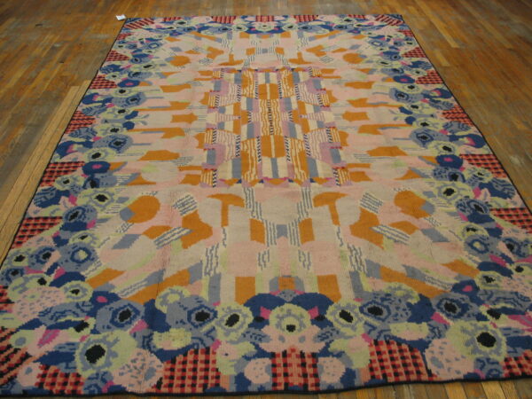 Abstract high-pile rug features pink, orange, and blue geometric patterns with a stylized floral border on wood planks.