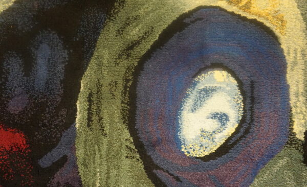 Textured close-up of a rug featuring an abstract eye-like pattern in blue, purple, olive green, and black.