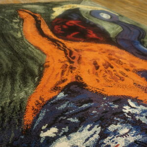 Plush abstract rug featuring bold orange, deep blue, and dark green shapes on light wood flooring.