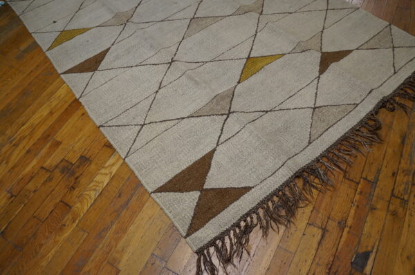 Beige flat-weave rug featuring geometric triangles in brown and gold, laid on worn hardwood flooring.