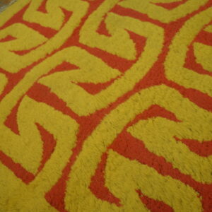 Close-up of a high-pile rug featuring a thick yellow and red interlocking geometric design on wood floorboards.