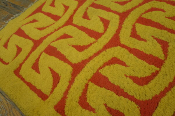 Close-up of a high-pile rug featuring a thick yellow and red interlocking geometric design on wood floorboards.