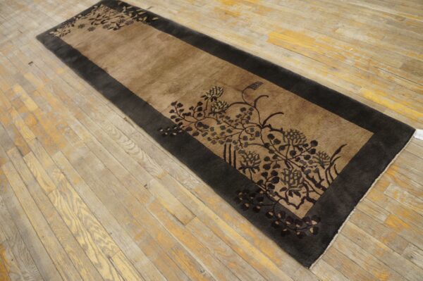 Tan and charcoal runner rug with a bold, dark floral motif lying on patchy wood planks.