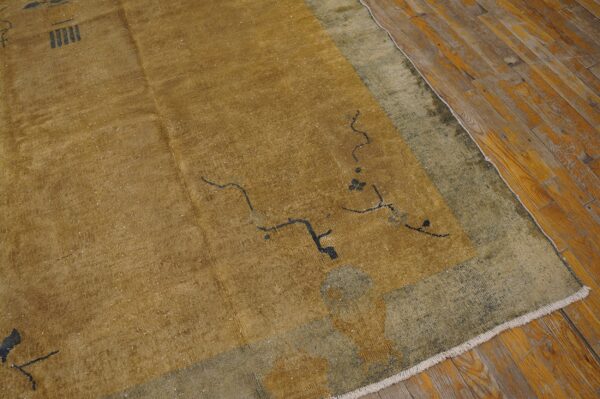 Low pile gold rug with sparse black designs and a distressed olive border on worn wood flooring.