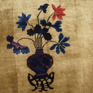 Lustrous tan rug featuring a central motif of a dark purple vase filled with indigo, magenta, and red flowers.