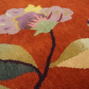 Stylized purple, green, and gold floral pattern on a textured deep rust orange rug surface.