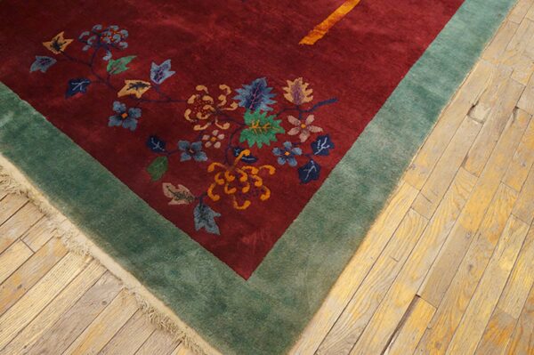 Burgundy rug with ornate floral pattern and sage green border rests on light wood planks.