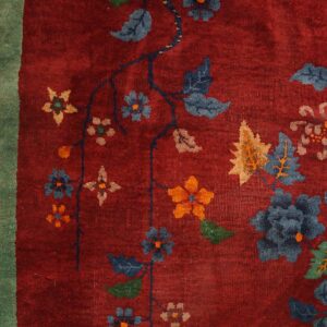 Detailed red rug featuring sprawling blue, orange, and tan floral designs bordered by green, resting on wood.