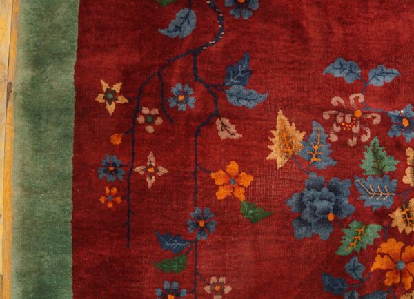 Detailed red rug featuring sprawling blue, orange, and tan floral designs bordered by green, resting on wood.