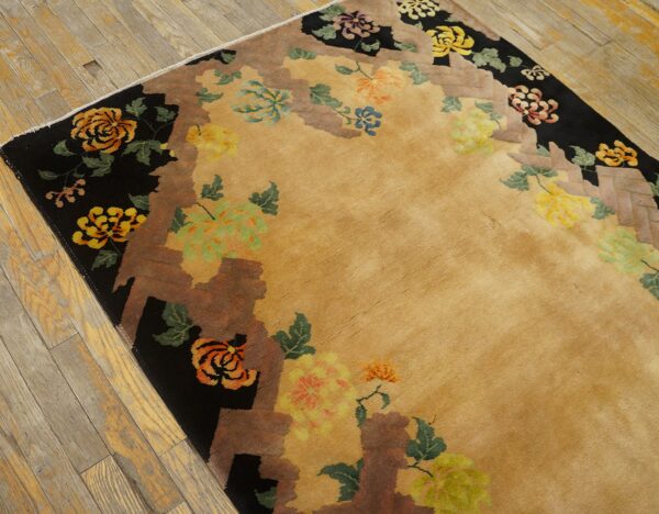Tan rug with colorful floral motifs, black and brown stepped edges, lies on worn hardwood flooring.