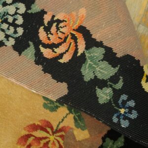 Detail of a rolled rug edge revealing tightly woven black and peach floral borders over a gold low-pile rug.