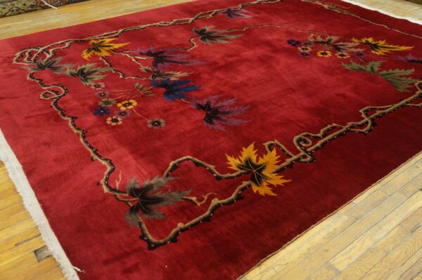 A deep red rug with a low pile featuring a thorny vine border with multicolored maple leaves and small flowers, displayed on wood floors.