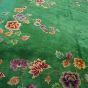 Deep green rug with large multicolored floral vines and a light teal border on wood flooring.