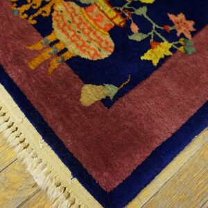 Plush rug corner with navy floral patterns, a maroon border, and fringe on yellow wood plank flooring.