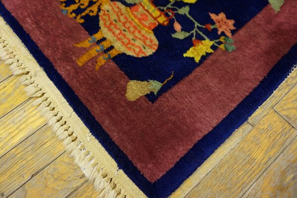 Plush rug corner with navy floral patterns, a maroon border, and fringe on yellow wood plank flooring.