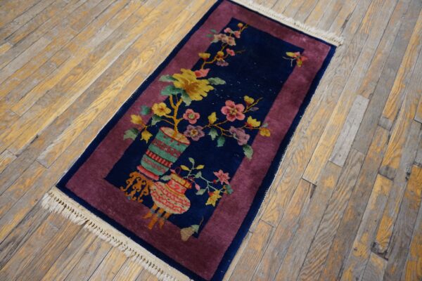 A small navy blue rug with a floral vase design and magenta border lies on aged wood floorboards.