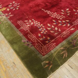 Red floral rug with a thick olive green border and geometric detailing rests on worn light wood flooring.