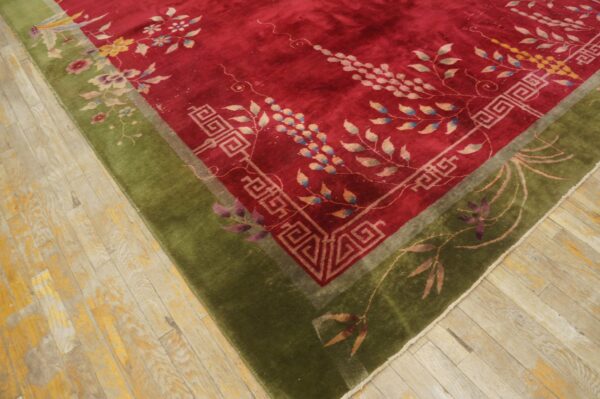 Red floral rug with a thick olive green border and geometric detailing rests on worn light wood flooring.