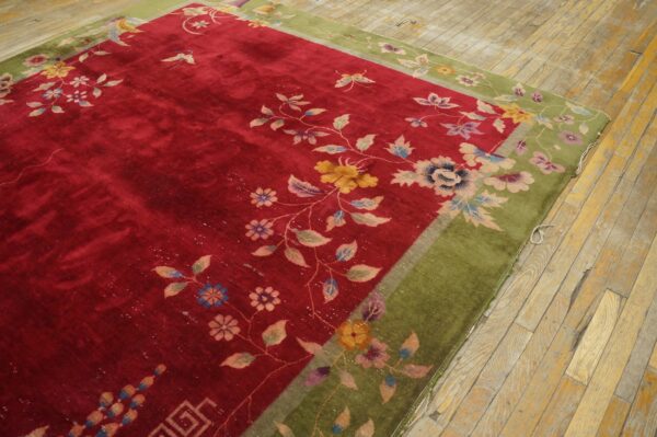 A velvety red floral rug with a moss green border rests diagonally on distressed wood flooring.