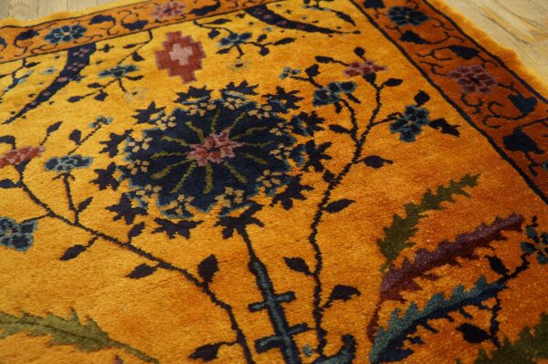 Close-up of a mustard yellow rug patterned with navy, teal, and maroon classical floral designs on wood floor.