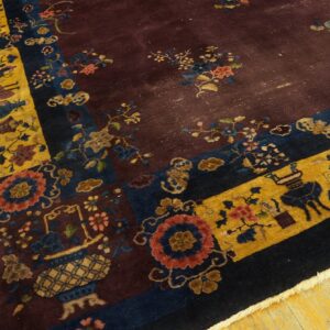 A low-pile burgundy rug with patterned gold, navy, and multi-colored borders rests on yellow wood flooring.